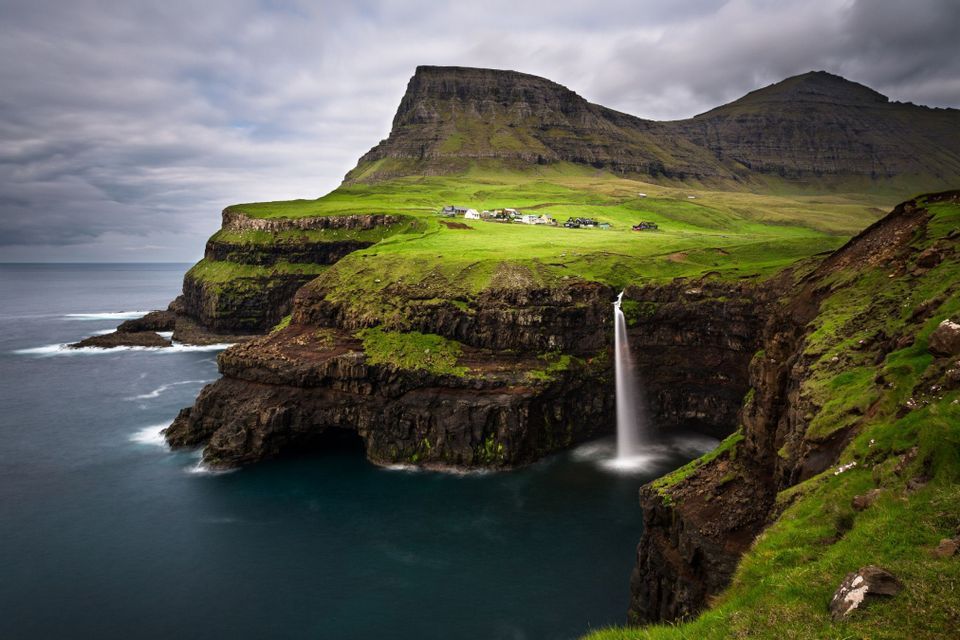Una cascata si riversa da una verde scogliera erbosa direttamente nell'oceano, con un piccolo villaggio incastonato sulla collina sullo sfondo.