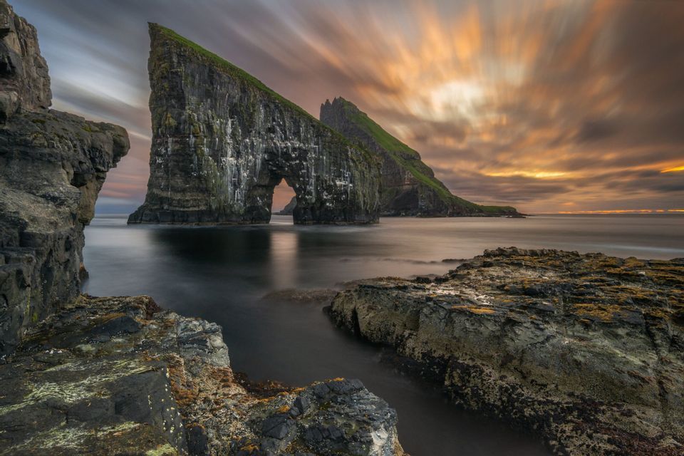 Scatto a lunga esposizione di due grandi faraglioni, uno con un arco naturale, su una costa rocciosa sotto un drammatico cielo al tramonto.