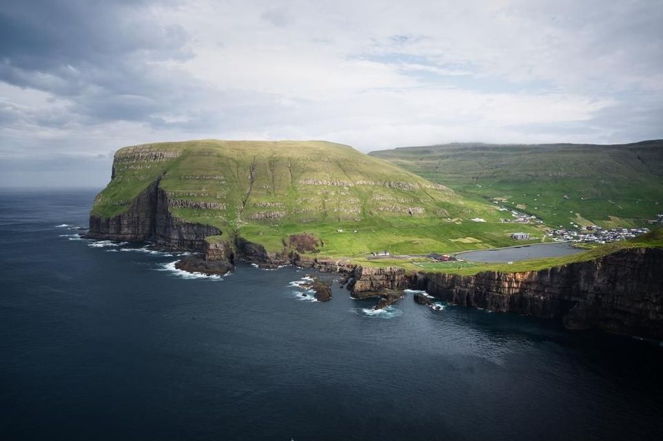Una vista aerea di una grande scogliera verdeggiante su una costa frastagliata, con un piccolo villaggio e un lago sullo sfondo sotto un cielo nuvoloso.