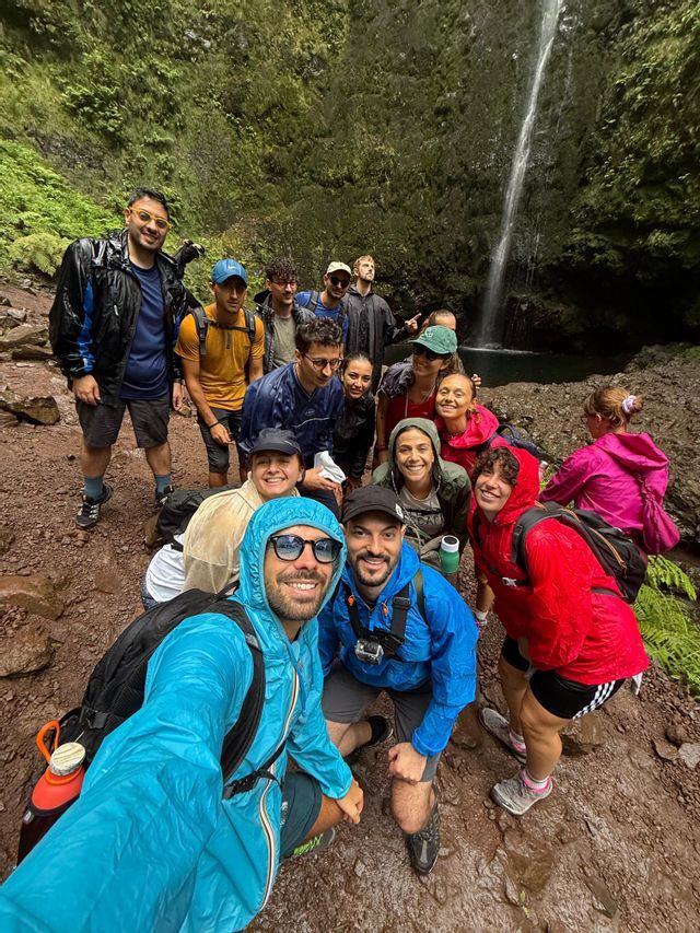 Un gruppo WeRoad in impermeabili colorati posa per un selfie davanti a un'alta cascata in una foresta lussureggiante.