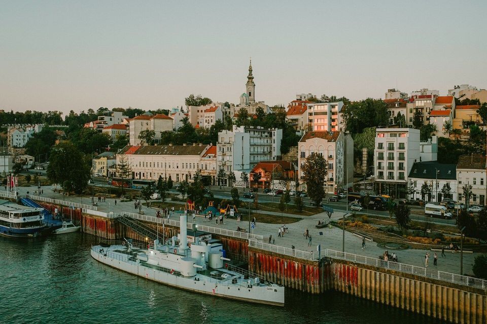 Ein weißes Schiff liegt an einem Stadtkai an einem Fluss, mit einer historischen Stadtlandschaft und einem Kirchturm im Hintergrund bei Sonnenuntergang.