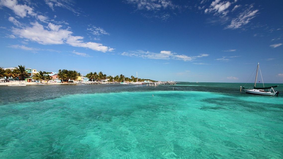Una vista sull'acqua limpida e turchese che mostra una costa tropicale con palme, edifici colorati e una barca a vela bianca ancorata.