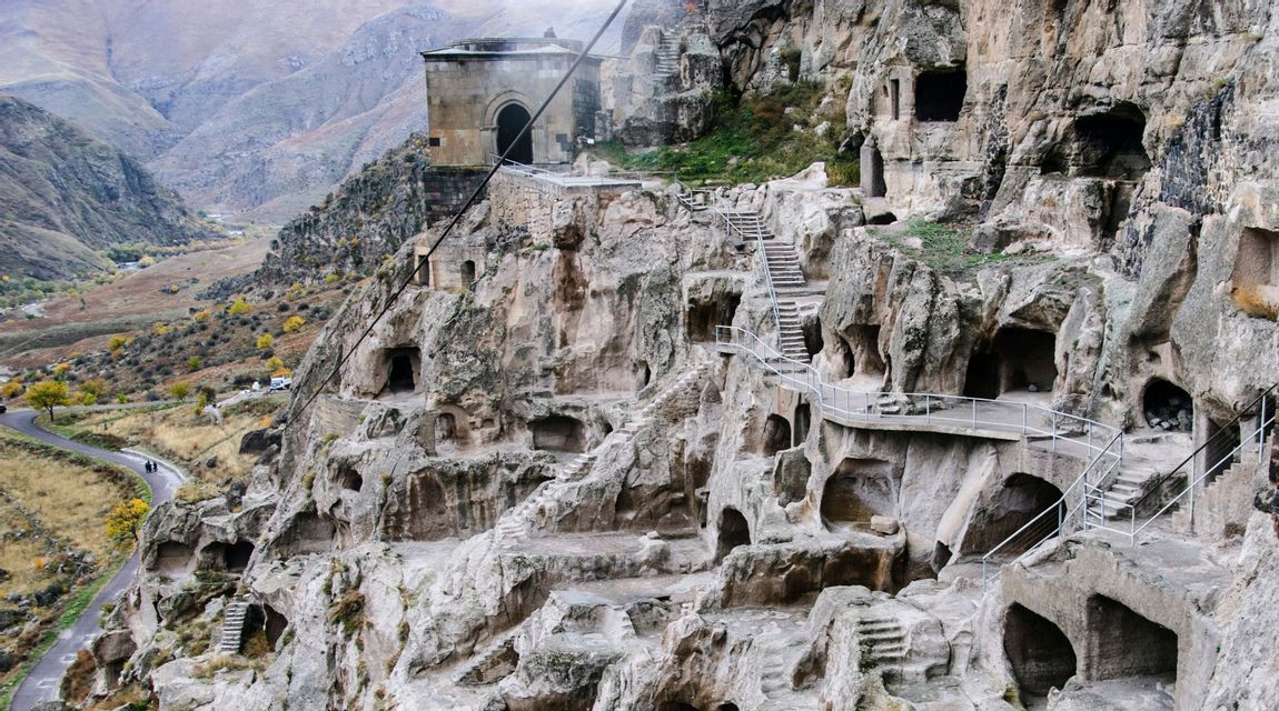 Un monastero rupestre a più livelli scavato in una parete rocciosa, con scalinate e passerelle che si affacciano su una valle montuosa.
