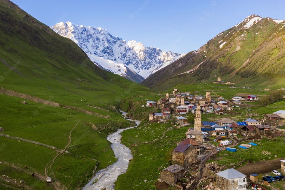 Una vista aerea di un piccolo villaggio con torri in pietra in una valle verde, situato vicino a un fiume con montagne innevate sullo sfondo.