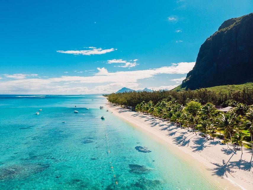 Vista aerea di una costa tropicale con spiaggia di sabbia bianca, palme, acqua turchese cristallina e una montagna verde.