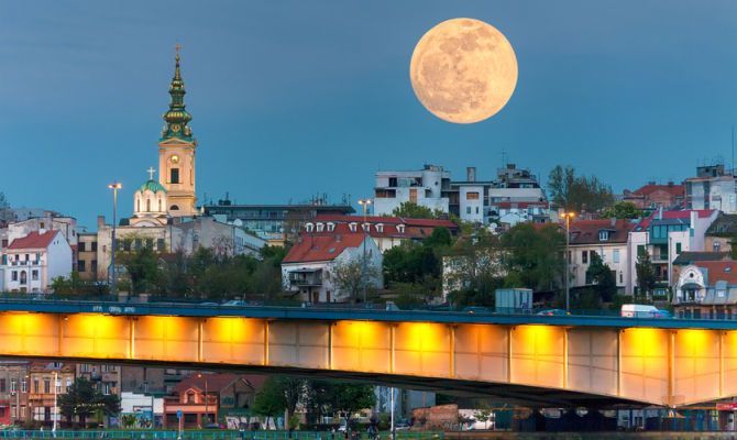 Une grande pleine lune se dresse dans le ciel crépusculaire au-dessus d'une silhouette urbaine, avec un clocher d'église et un pont illuminé au premier plan.