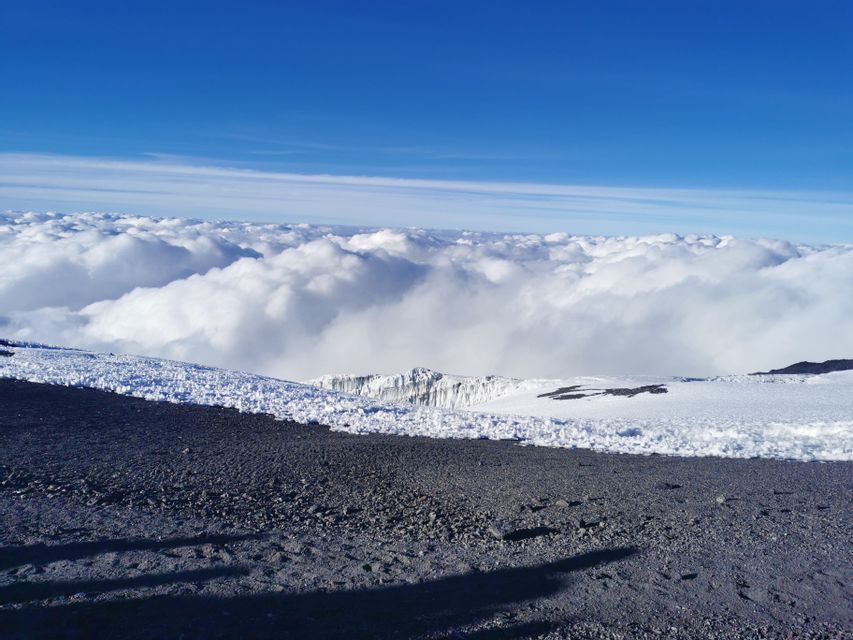 Una vista da una cima rocciosa e innevata, che guarda su un mare di nuvole bianche sotto un cielo azzurro limpido.