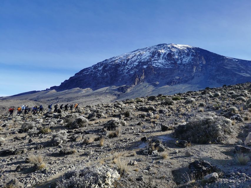Un gruppo WeRoad in viaggio fa trekking in fila attraverso un paesaggio roccioso e d'alta quota, con un'imponente montagna innevata in lontananza.