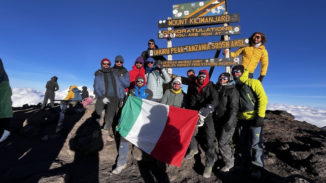 Un gruppo WeRoad in viaggio posa con la bandiera italiana sulla vetta Uhuru Peak del Monte Kilimanjaro, sopra un mare di nuvole.
