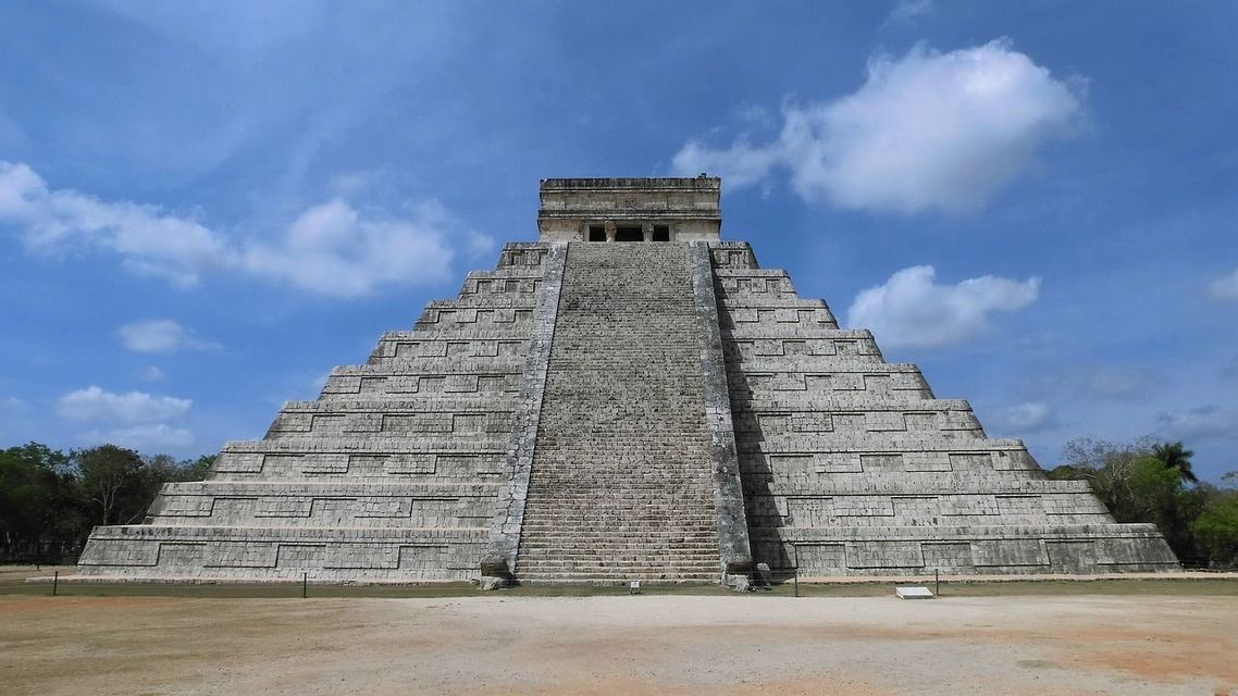 Una grande piramide a gradoni in pietra, con una scalinata centrale, si staglia al centro dell'immagine sotto un cielo blu con nuvole bianche.