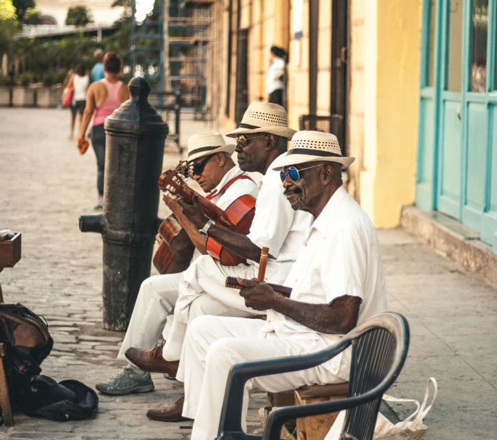 Three musicians in white outfits and hats sit on a cobblestone street playing guitars and other instruments.
