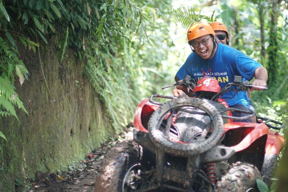 Two people on a WeRoad group trip laughing while riding a muddy red quad bike on a trail through a lush jungle.