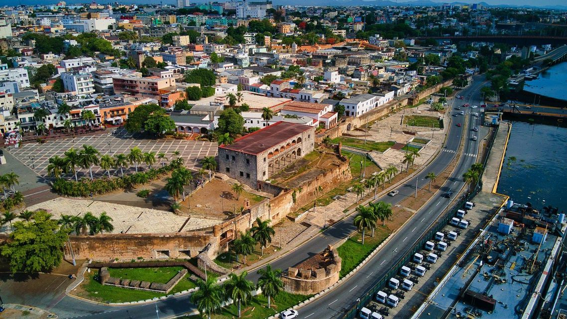 An aerial view of a historic coastal city showing ancient stone fortifications, a large plaza, and a modern road alongside the water.