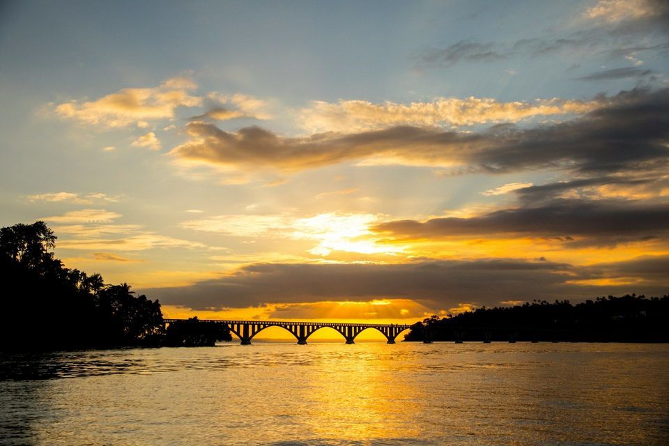 A silhouetted arched bridge crosses a body of water as the sun sets, casting a golden glow on the clouds and water.