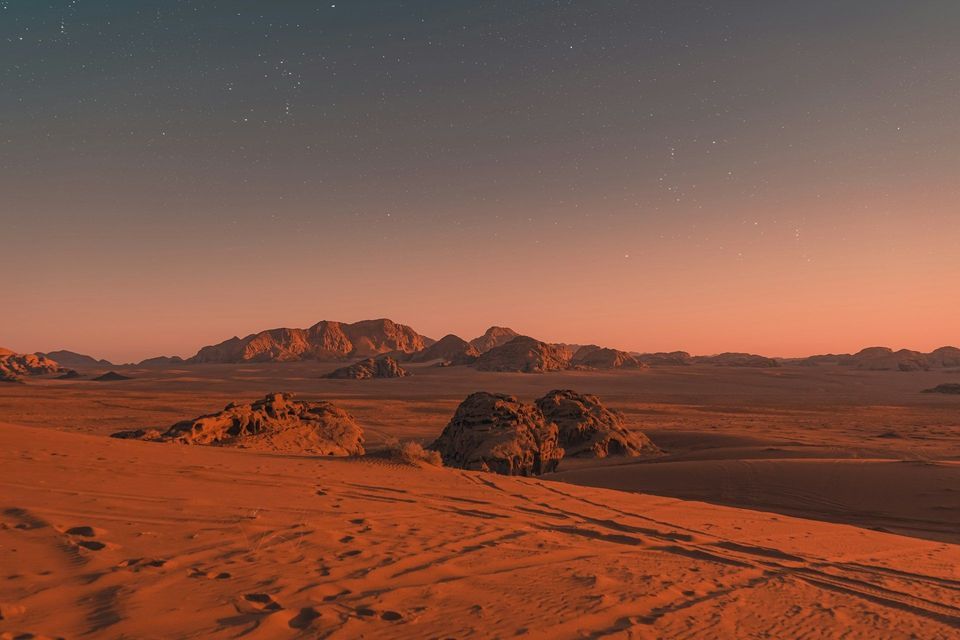 A vast desert landscape with red sand and rocky mountains under a starry sky at dusk.