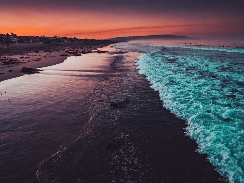 Turquoise waves roll onto a dark sand beach, reflecting the vibrant orange and purple colors of the sunset sky.