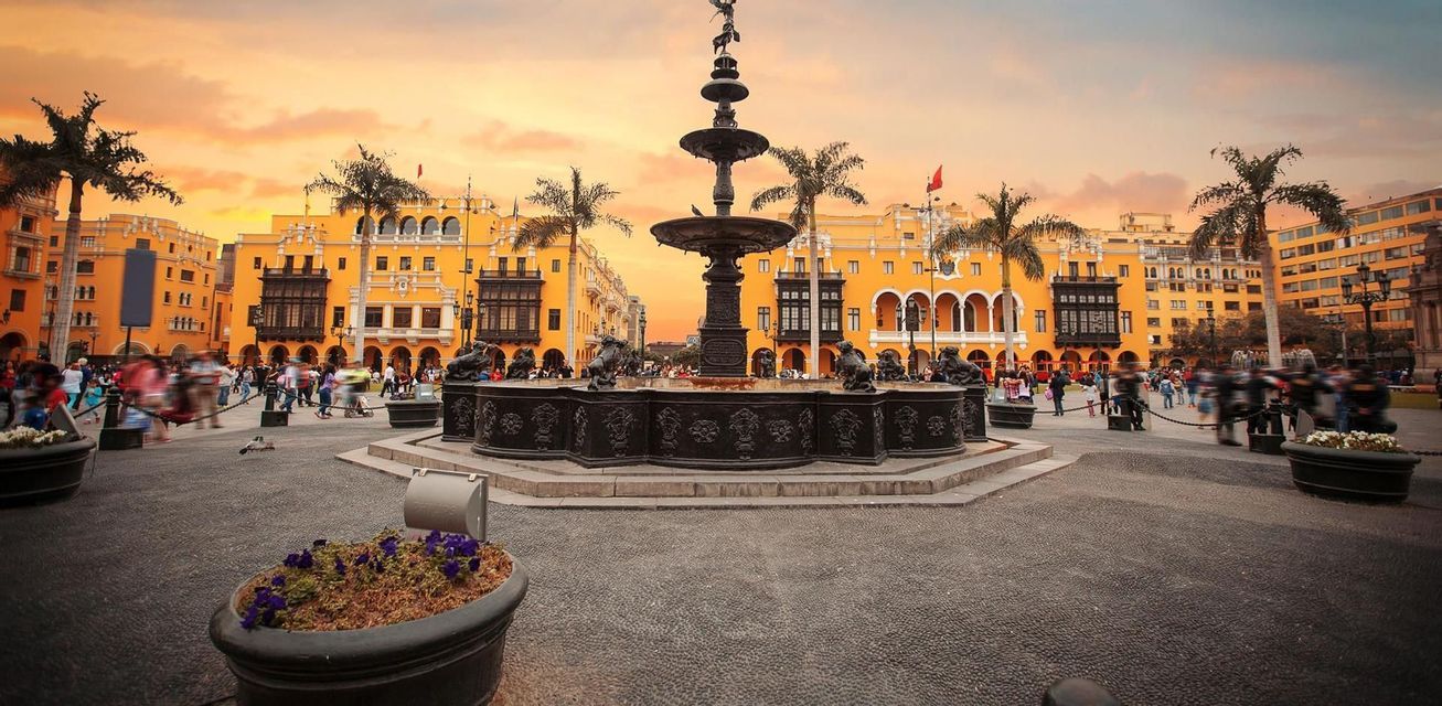 An ornate fountain stands in the center of a city plaza surrounded by yellow colonial-style buildings and palm trees at sunset.