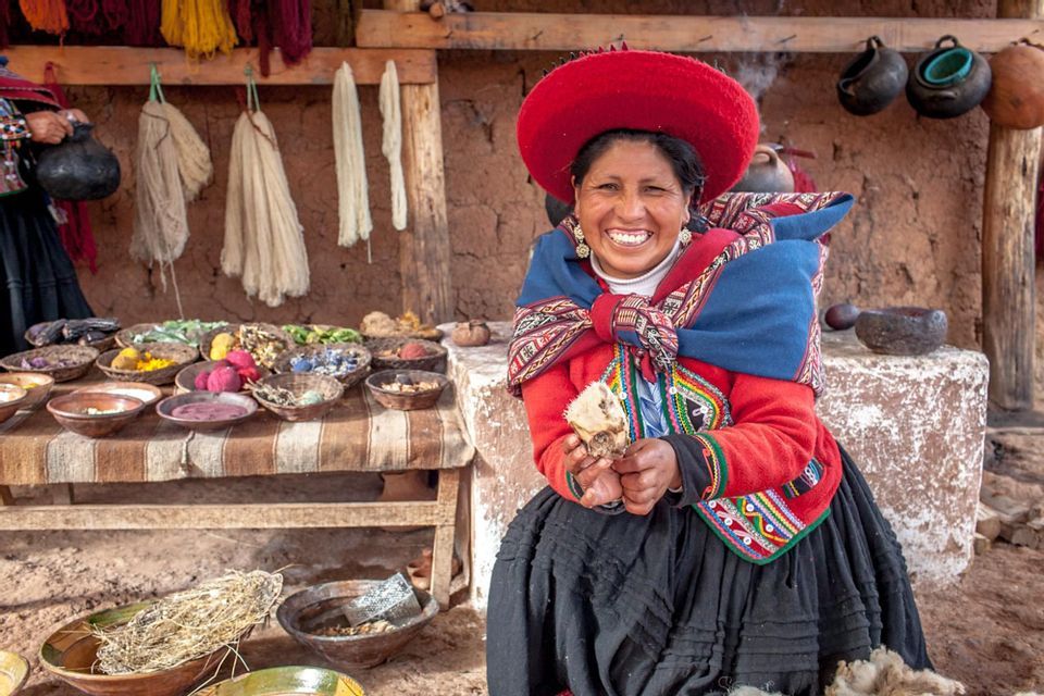 A smiling woman in traditional attire sits at a textile workshop, holding raw wool, with bowls of natural dyes on a table beside her.