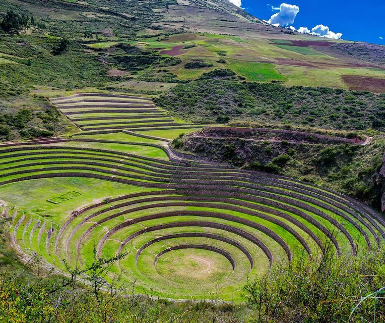 Concentric circular agricultural terraces with stone walls built into a lush green hillside under a blue sky.