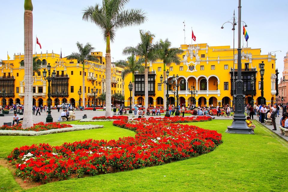 A bustling city square with a large yellow colonial building, palm trees, and vibrant red flowerbeds on a green lawn where people gather.