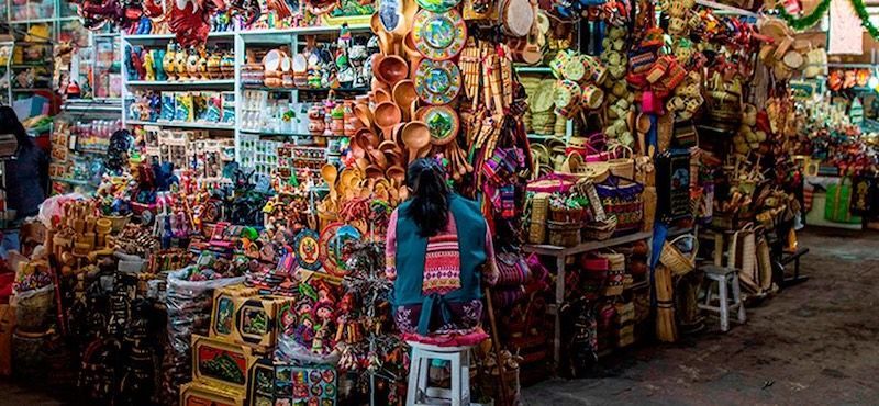 A woman with her back to the camera sits on a stool in a busy market stall filled with colorful handmade souvenirs and crafts.