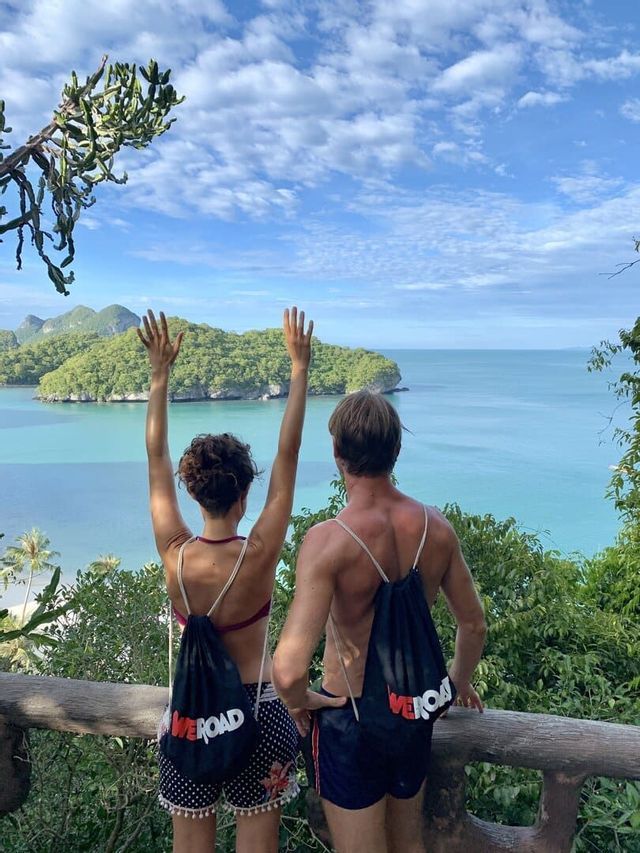 A man and a woman on a WeRoad group trip admire a tropical bay with green islands, the woman raises her arms in the air.