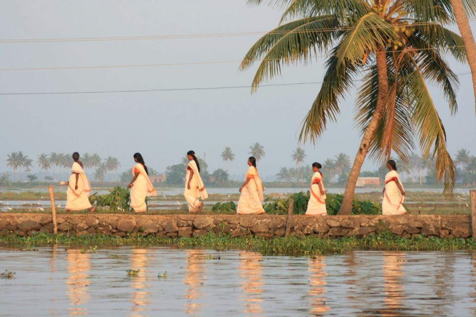 Sei donne in tradizionali sari bianchi camminano in fila indiana su un sentiero di pietra lungo un fiume con palme.