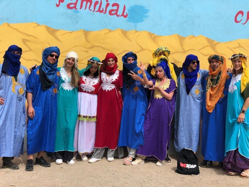 Un groupe WeRoad, vêtu de robes traditionnelles colorées et de foulards, pose pour une photo devant un mur de désert peint.