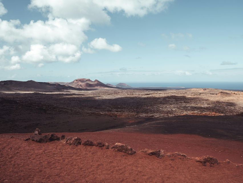 Un vasto paesaggio vulcanico con terreno rosso in primo piano si estende verso montagne distanti e l'oceano sotto un cielo nuvoloso.