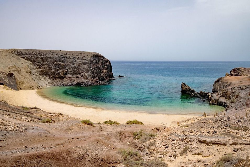Panoramica da una scogliera rocciosa su una spiaggia sabbiosa appartata, incastonata in una caletta con acque turchesi e cristalline, sotto un cielo leggermente velato.