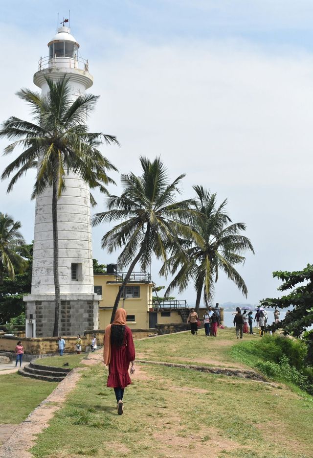 Una persona con un vestito rosso e un foulard sale su una collina erbosa verso un faro bianco circondato da palme.