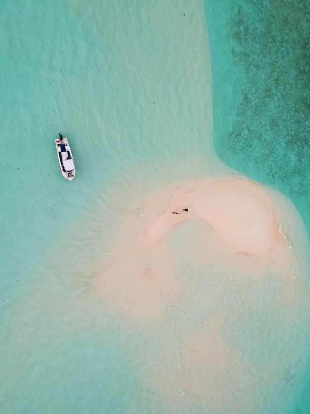 Vista aerea di una piccola barca bianca che galleggia in acqua turchese cristallina, vicino a un banco di sabbia con due persone.