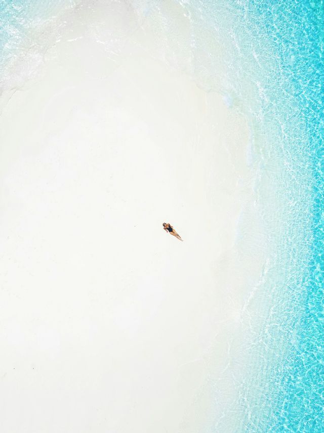 Vista aerea di una persona in costume da bagno sdraiata su una spiaggia di sabbia bianca accanto ad acqua turchese cristallina.