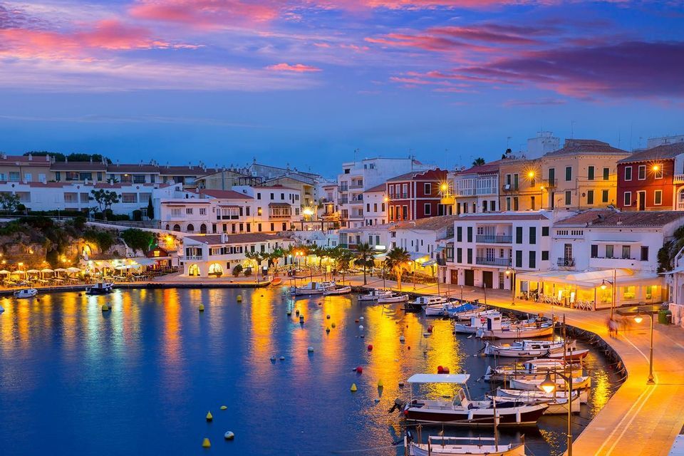 A coastal town's harbor illuminated at dusk, with lights from buildings reflecting on the water where boats are moored.