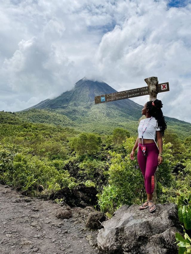 A woman on a hiking trail stands on a rock, looking towards a large volcano partially covered by clouds.