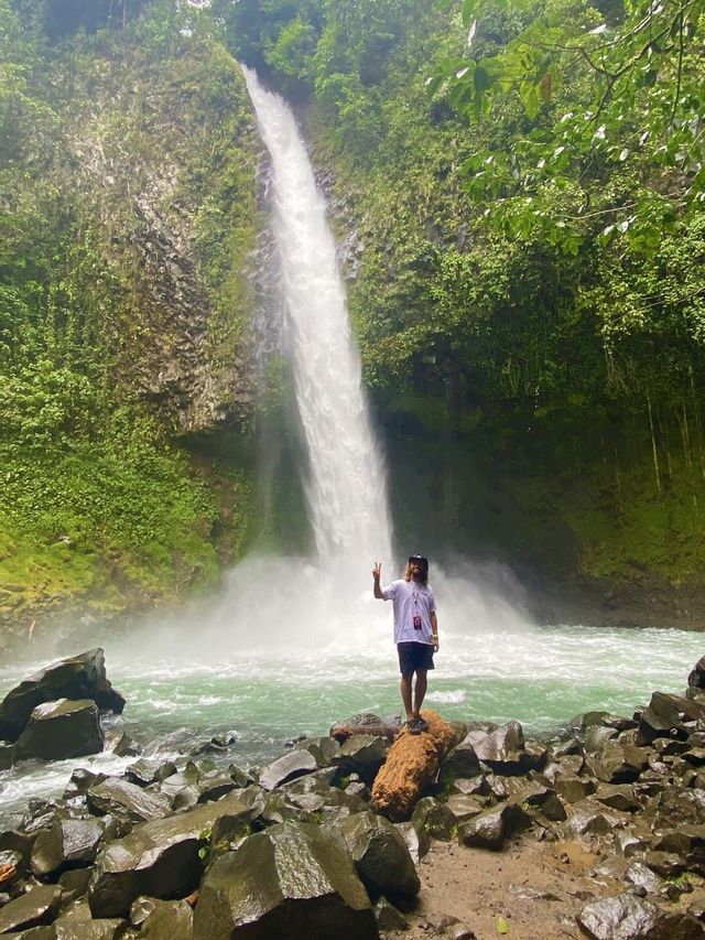 Eine Person mit langen Haaren steht auf einem Felsen und macht ein Friedenszeichen vor einem hohen Wasserfall in einem üppig grünen Dschungel.