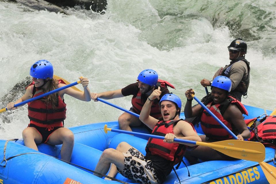 A WeRoad group trip smiles while rafting on whitewater rapids in a blue boat, wearing helmets and life vests.