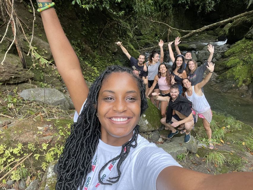 Una donna sorridente con i dreadlock si scatta un selfie con il suo gruppo di viaggio WeRoad mentre posano accanto a un ruscello roccioso in una foresta lussureggiante.