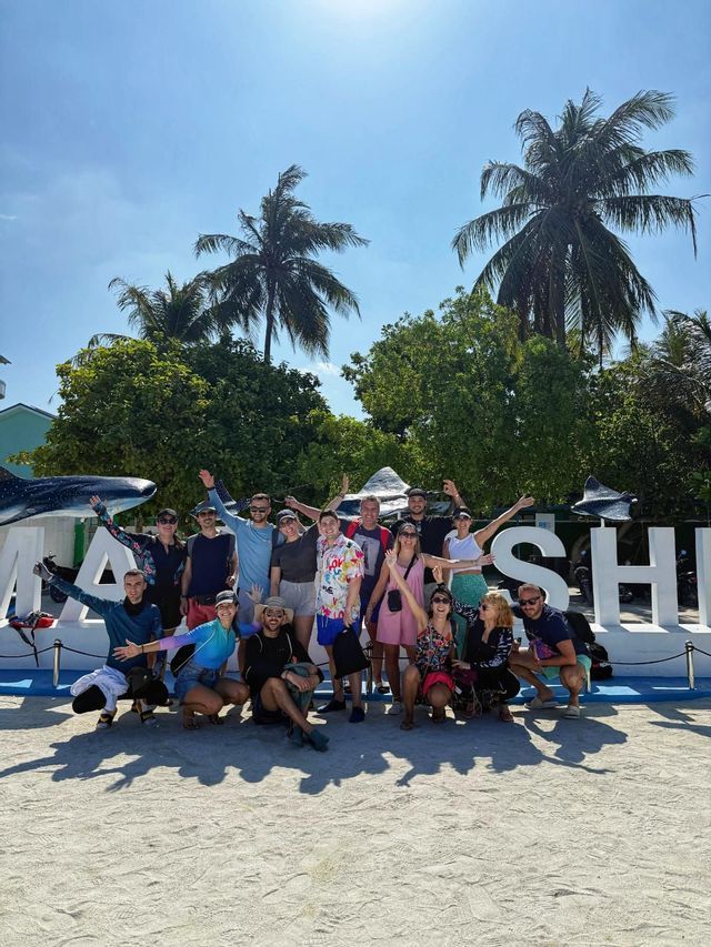 Un grupo de WeRoad posa para una foto en una playa de arena frente a un gran letrero con palmeras de fondo.