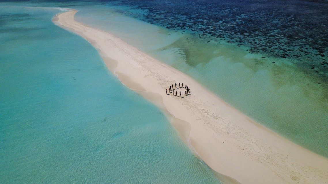 Vista aérea de un grupo de viaje de WeRoad de pie en círculo sobre un estrecho banco de arena blanca rodeado de agua turquesa.