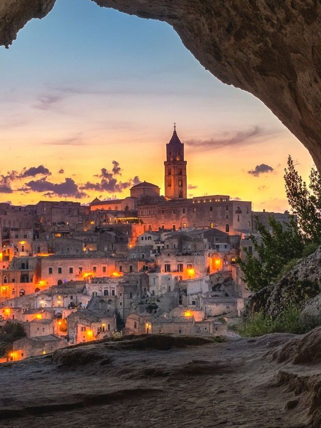 Un'antica città collinare con un campanile imponente, illuminata da luci calde al tramonto e vista dall'interno di una grotta.