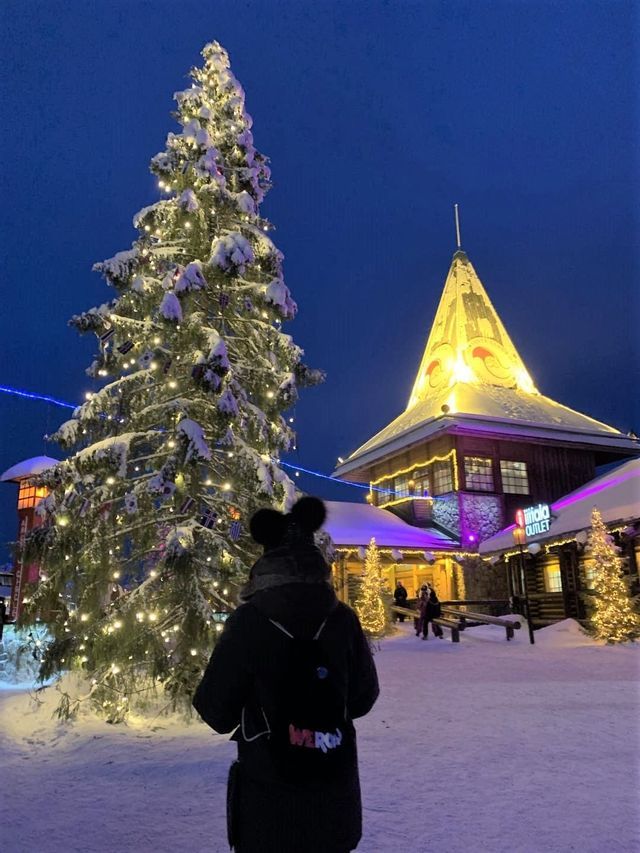 A person wearing a WeRoad backpack looks at a large, snow-covered Christmas tree illuminated with lights in a village square at night.