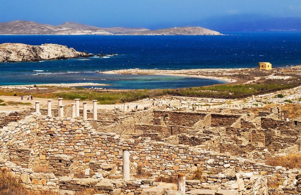 Antiche rovine in pietra con colonne bianche che si affacciano su un mare blu profondo e isole distanti sotto un cielo sereno.