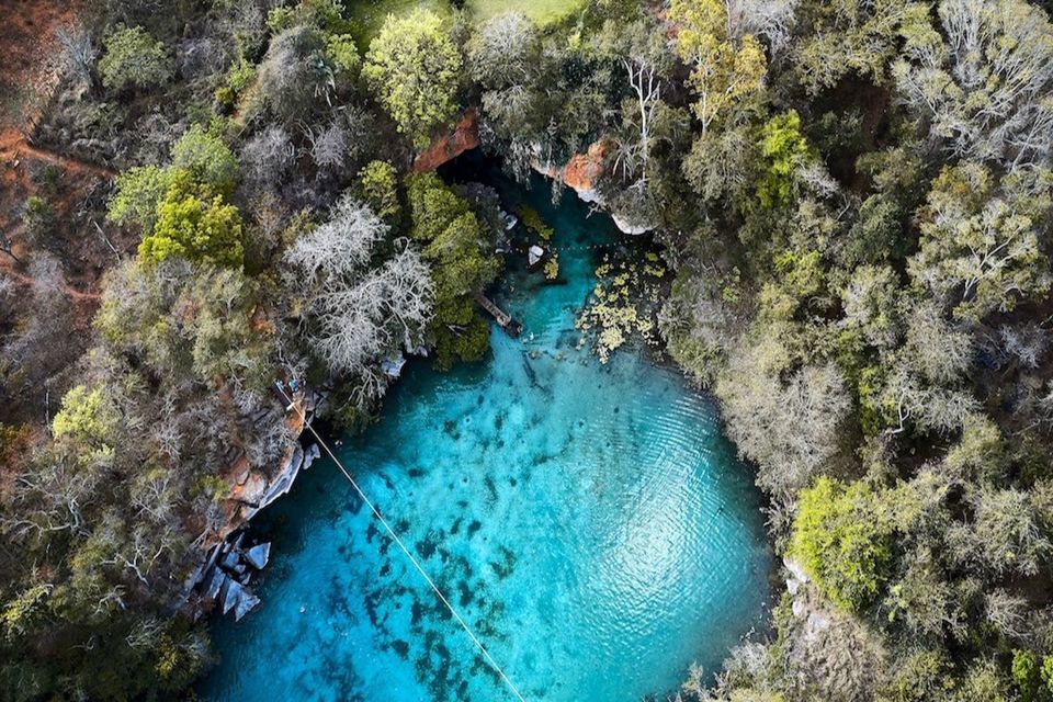 Una vista aerea di un cenote turchese circondato da una fitta foresta, con persone che nuotano nell'acqua cristallina sottostante.