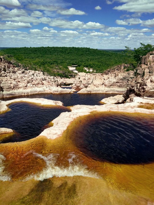Una vista panoramica di piscine naturali di acqua scura all'interno di un paesaggio roccioso, con una fitta foresta verde sotto un cielo blu.