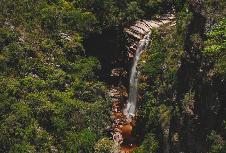 Una vista dall'alto di una cascata che si getta da una scogliera rocciosa in un laghetto rossastro, circondata da una lussureggiante foresta verde con persone radunate.