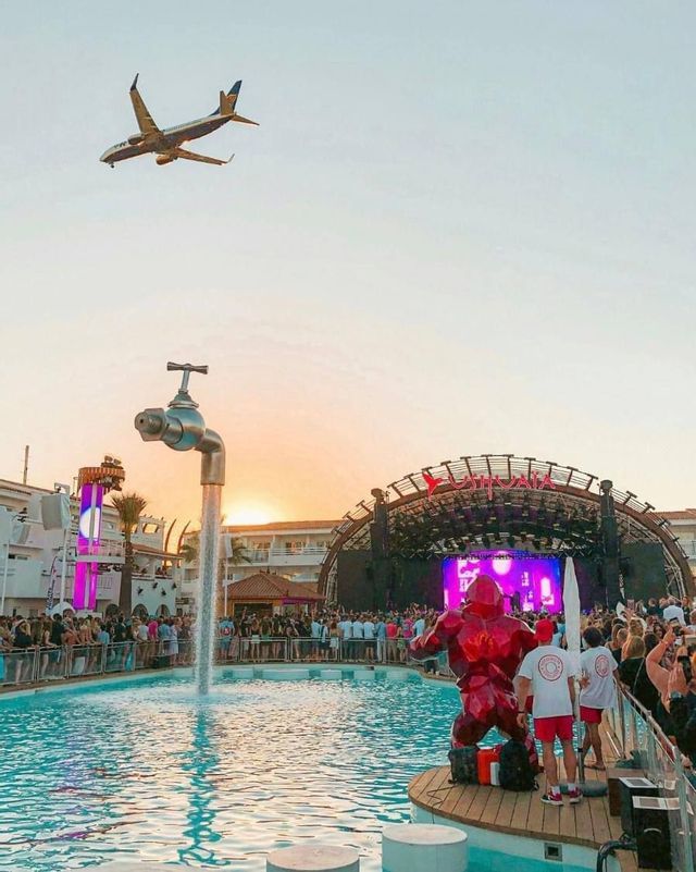 Un pubblico a un festival in piscina osserva un palco al tramonto, con una scultura di rubinetto galleggiante e un aereo a bassa quota.