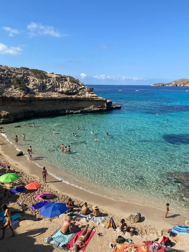 Vista dall'alto di una spiaggia in una cala, con persone che nuotano in acque turchesi cristalline e si rilassano sulla sabbia.