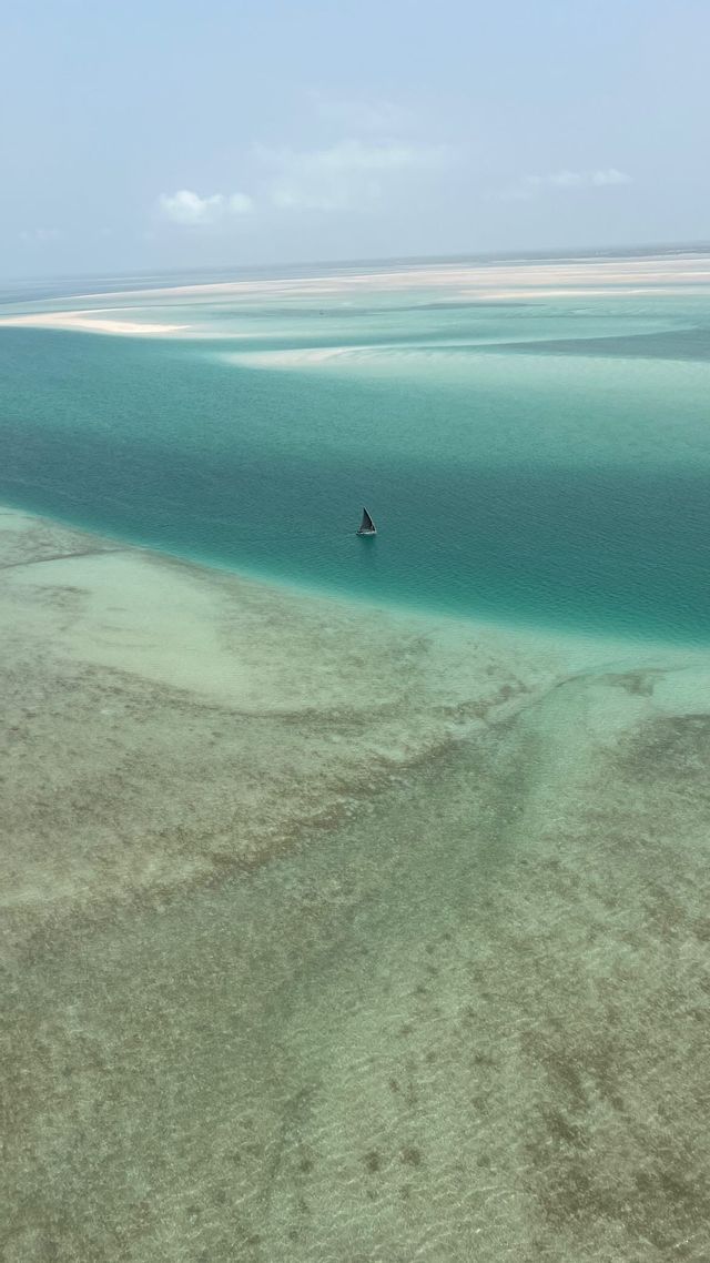 Vista aerea di una singola barca a vela su vaste acque turchesi e poco profonde con banchi di sabbia in lontananza.