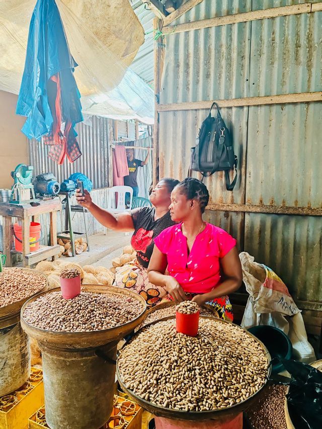 Una donna si fa un selfie con un'altra donna a una bancarella del mercato che vende grandi cesti di arachidi.